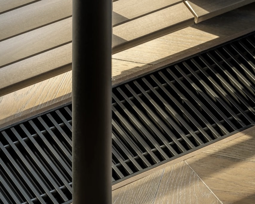 Крупний план дерев'яної підлоги з візерунком "ялинкою". У підлогу вмонтовано чорну вентиляційну решітку для опалення. Поруч — вертикальна чорна труба.