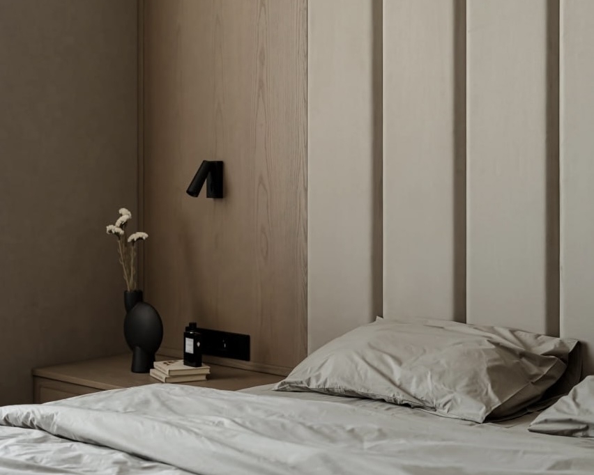 Інтер'єр спальні в спокійних бежевих тонах. Стіна за ліжком оздоблена м'якими панелями. Поруч — приліжкова тумба з вазою та настінний світильник.